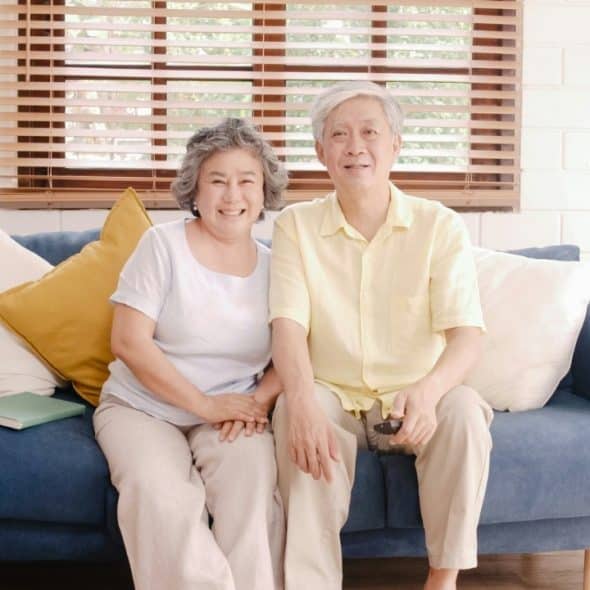 An older man and woman sitting on a blue sofa, smiling and holding a remote.