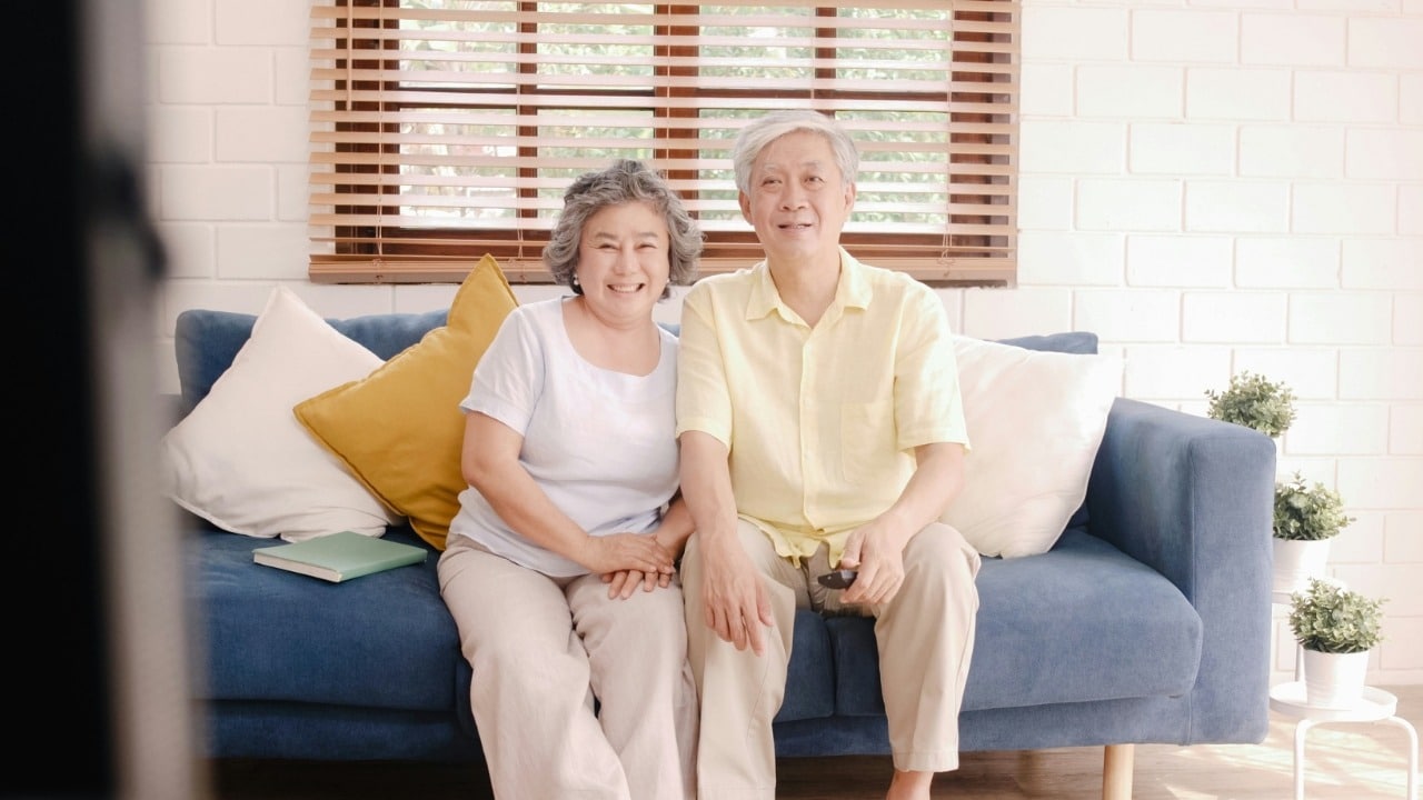 An older man and woman sitting on a blue sofa, smiling and holding a remote.