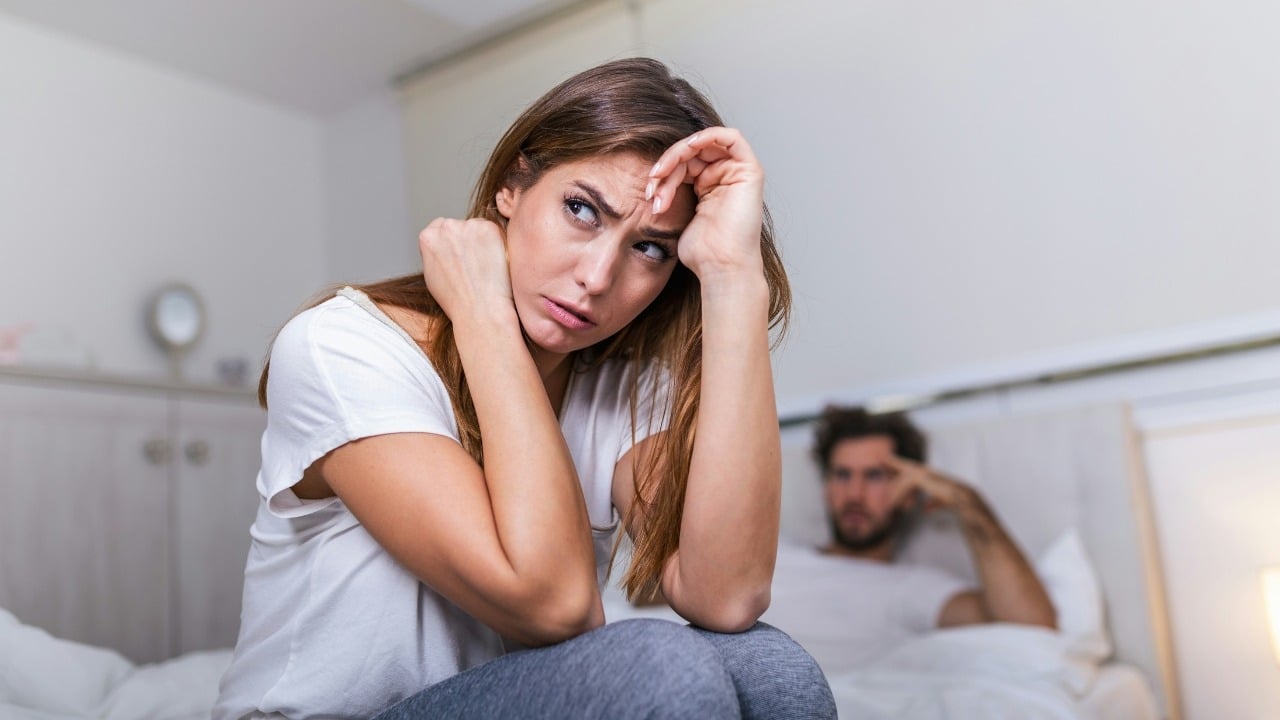 A woman sitting on a bed looking stressed while a man sits in the background.