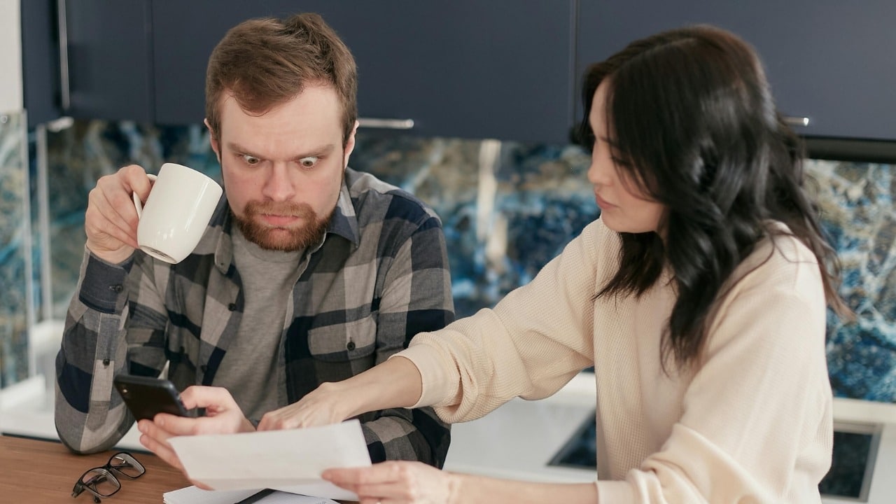 A man with a surprised expression looking at a phone while a woman holds papers.