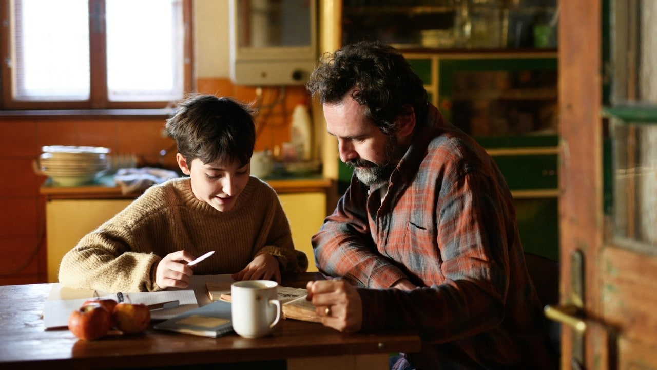 A bearded man and a young person sitting at a table looking at a notebook.