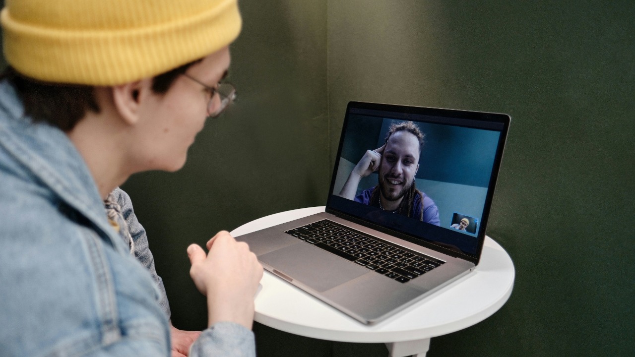 A person wearing a yellow beanie video chatting with a smiling man on a laptop.