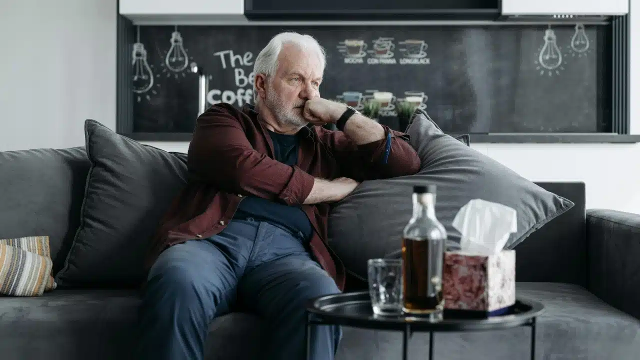 An older man sitting alone on a sofa, leaning his head on his hand.
