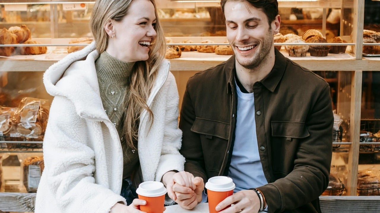A man and woman sitting outside a bakery, laughing and holding orange coffee cups.