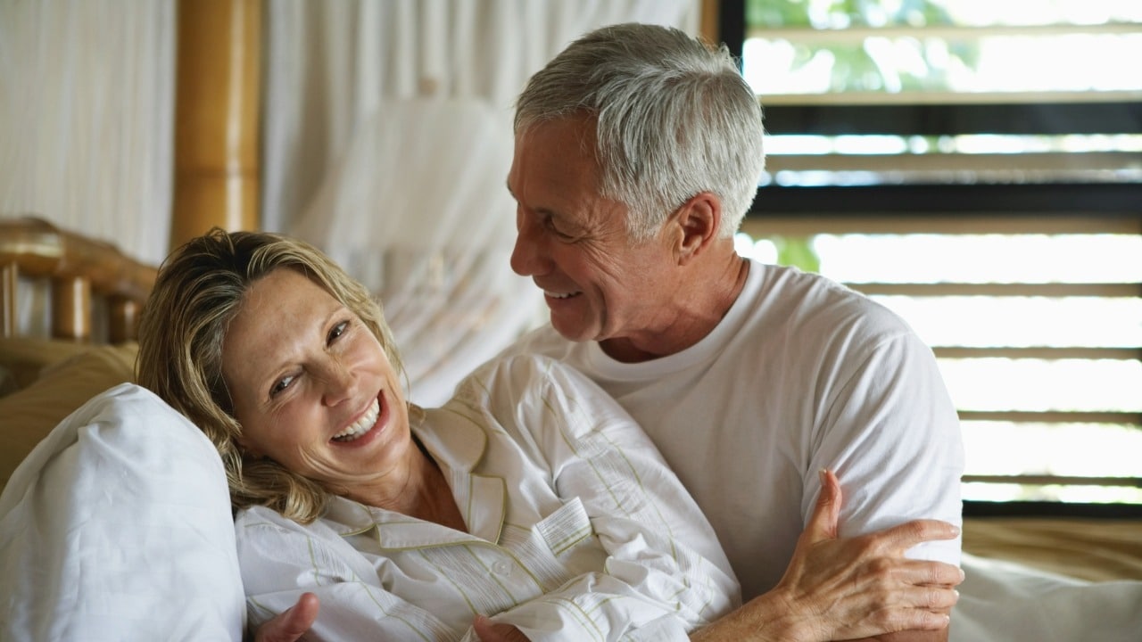 An older man and woman lying in bed, smiling and looking at each other.