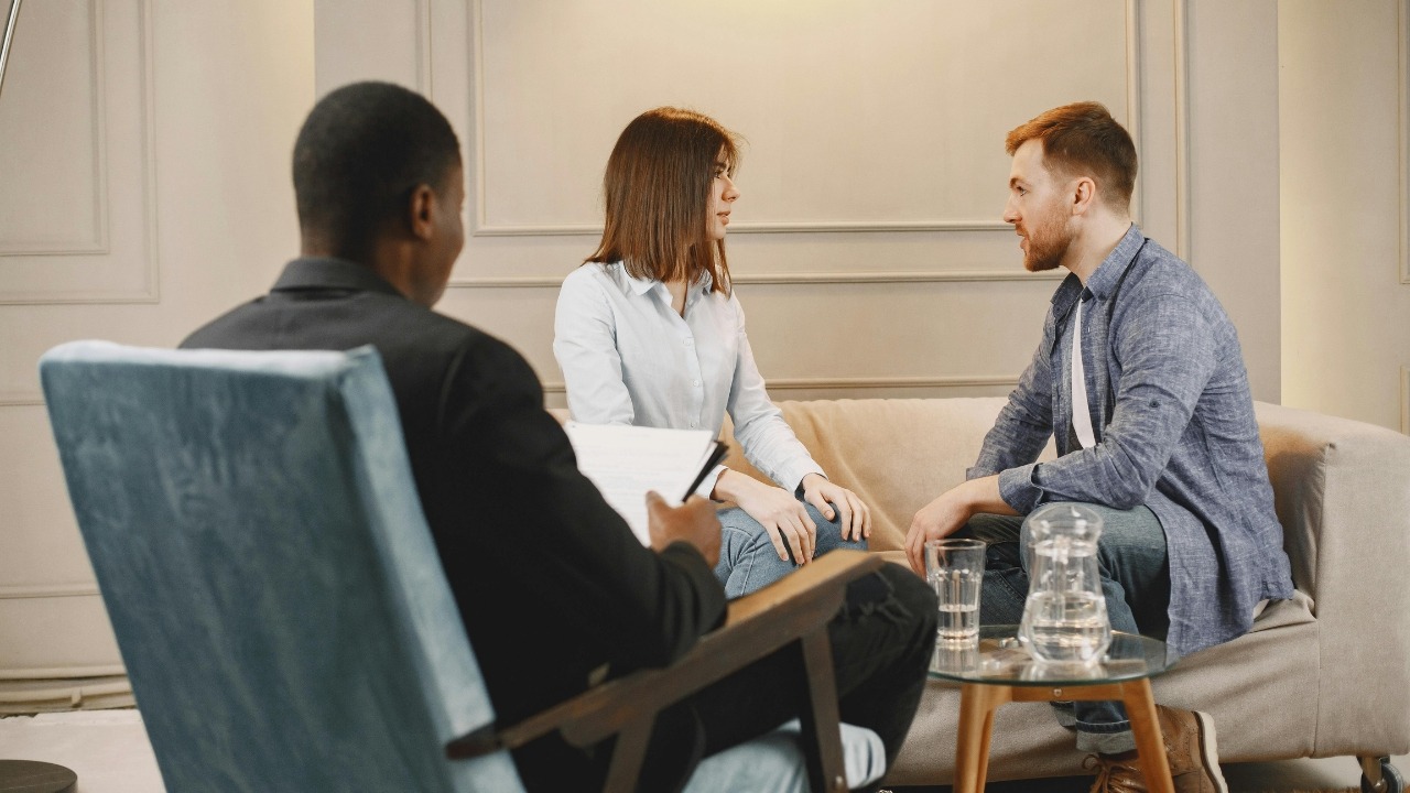 A man and woman sitting on a sofa talking to a person holding a clipboard.