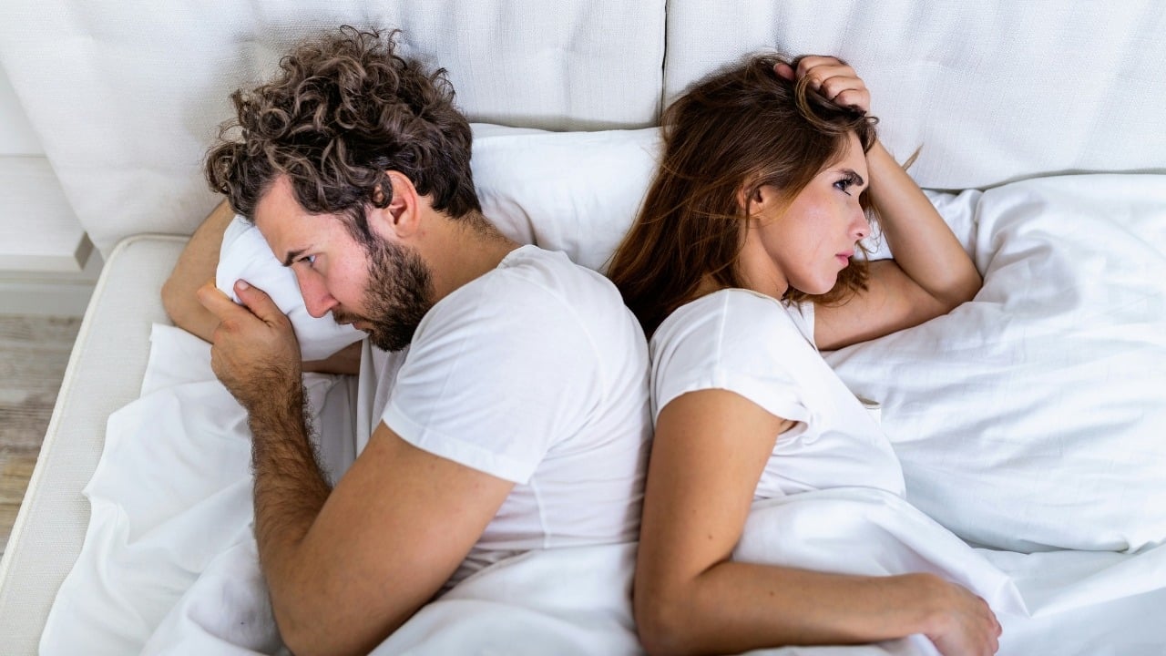 A man and woman lying in bed back-to-back with neutral expressions on white pillows.