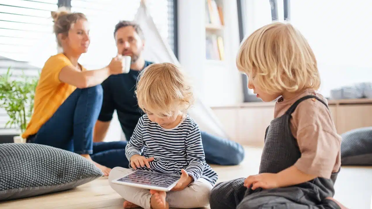 Two young children playing on the floor while two adults sit in the blurry background.