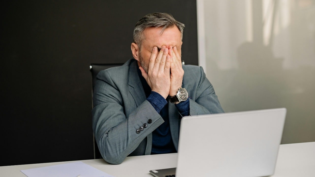 A man in a gray suit sitting at a desk with his hands over his face.