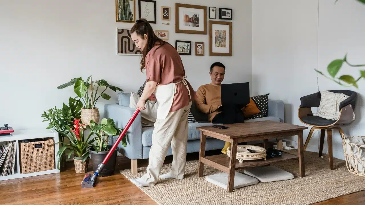 A woman vacuums a rug while a man sits on a sofa with a laptop.