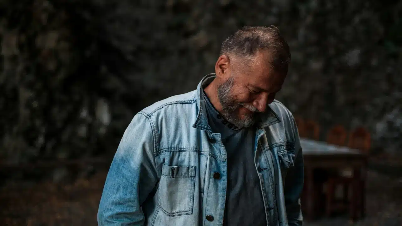A man with a beard wearing a denim jacket looks down and smiles in shadows.