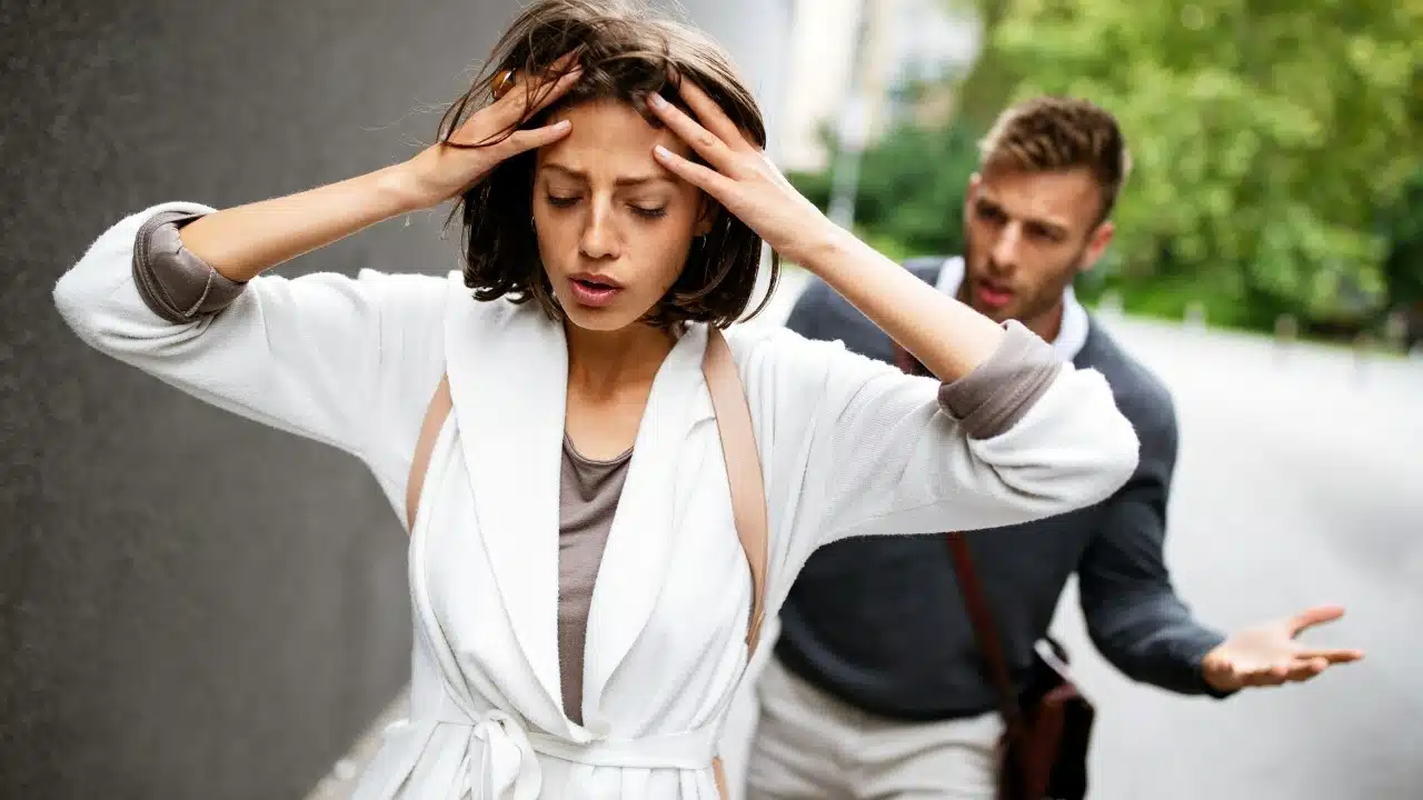A woman holds her head in her hands while a man gestures in the background.