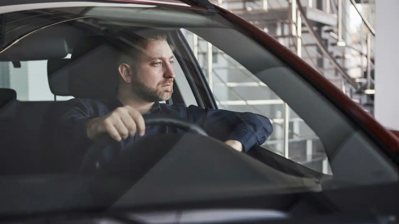 A man with a beard looks out the window while holding a car steering wheel.
