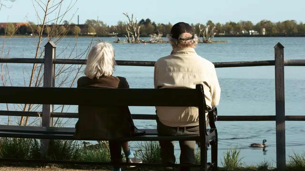 A senior man and woman sit on a bench looking out at a large lake.