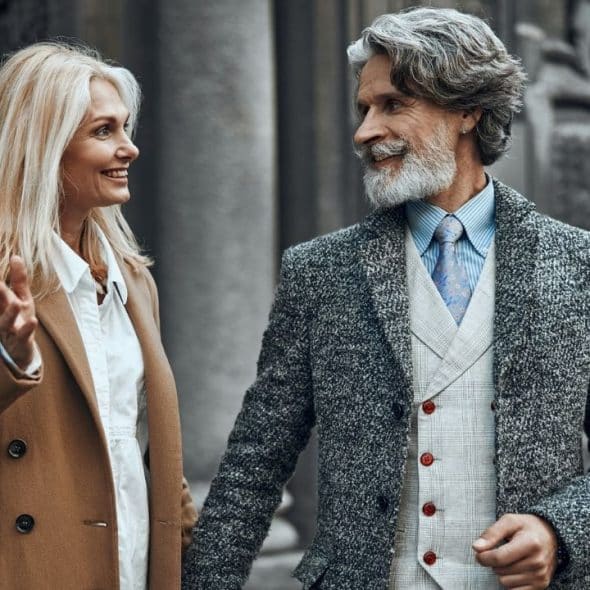 A smiling blonde woman in a tan coat talks to a bearded man in grey.