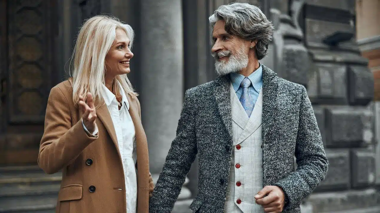 A smiling blonde woman in a tan coat talks to a bearded man in grey.