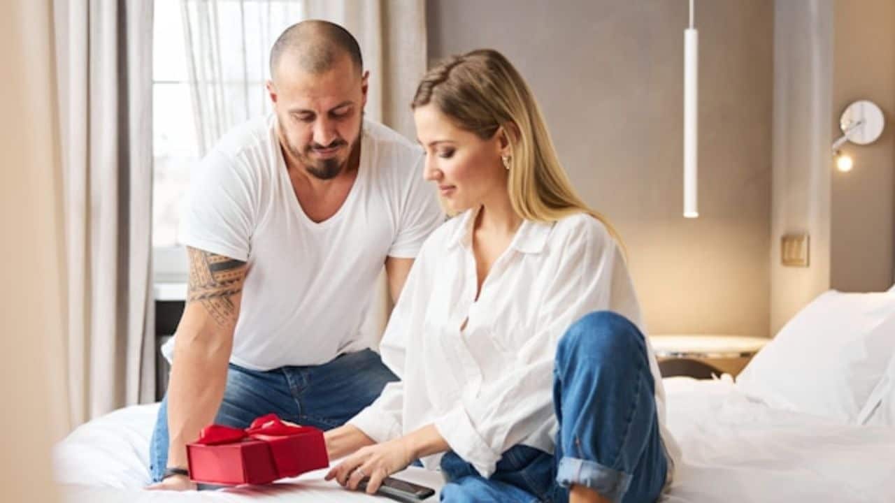 A woman gives a red gift box to a man with a tattooed arm.