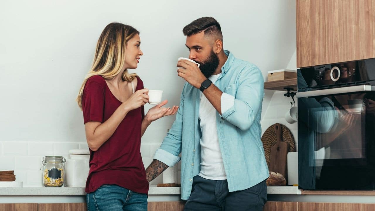 A woman in a maroon shirt and a bearded man drink from white mugs.