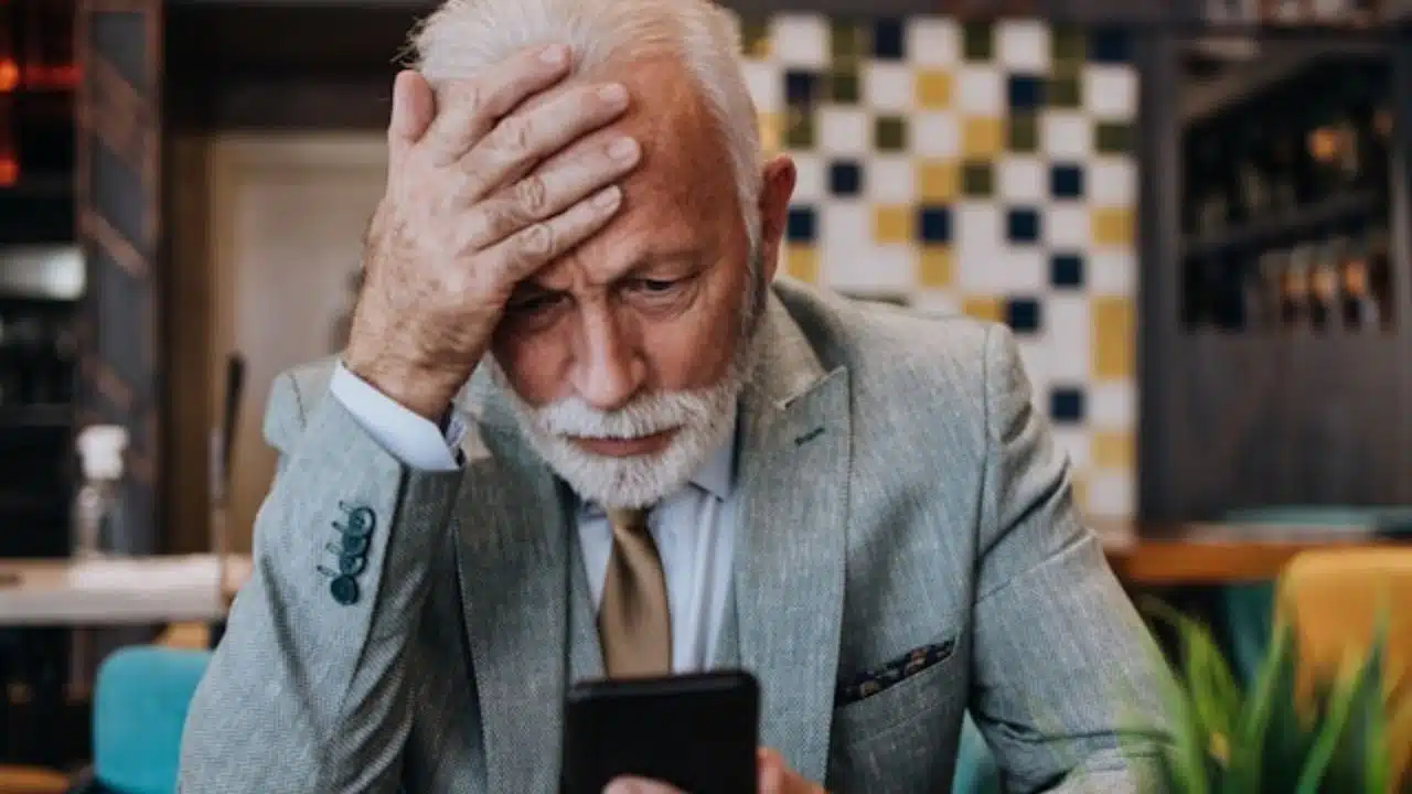 An older man in a suit holds his forehead while looking at a smartphone.