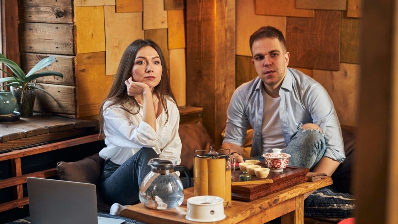 A man and woman sit behind a wooden table with a tea set and laptop.