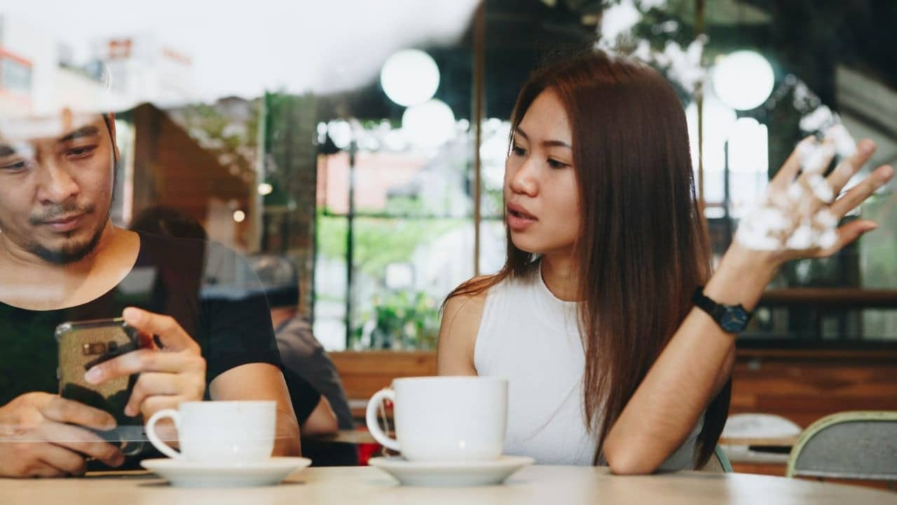 A woman talks and gestures while a man next to her looks at a phone.