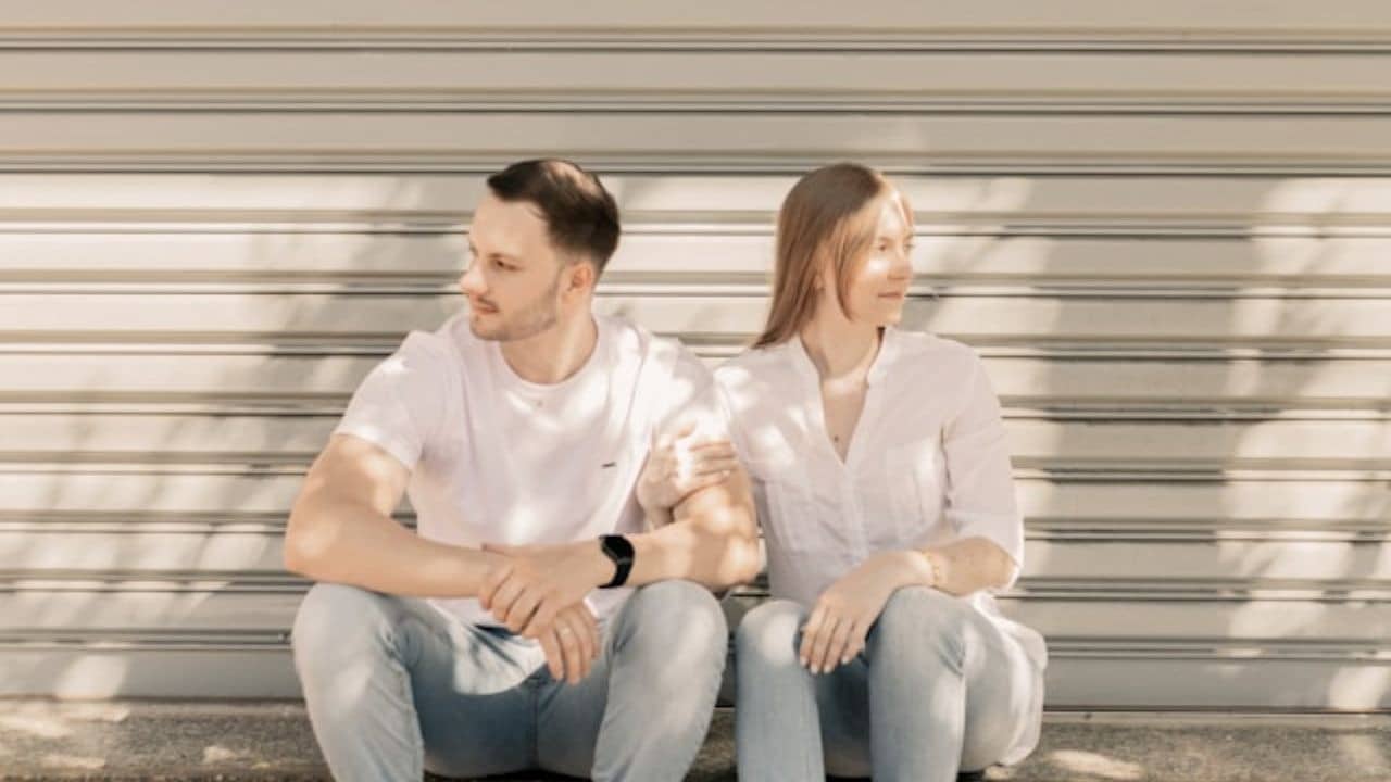 A man and woman sit in front of a metal shutter, both looking away.