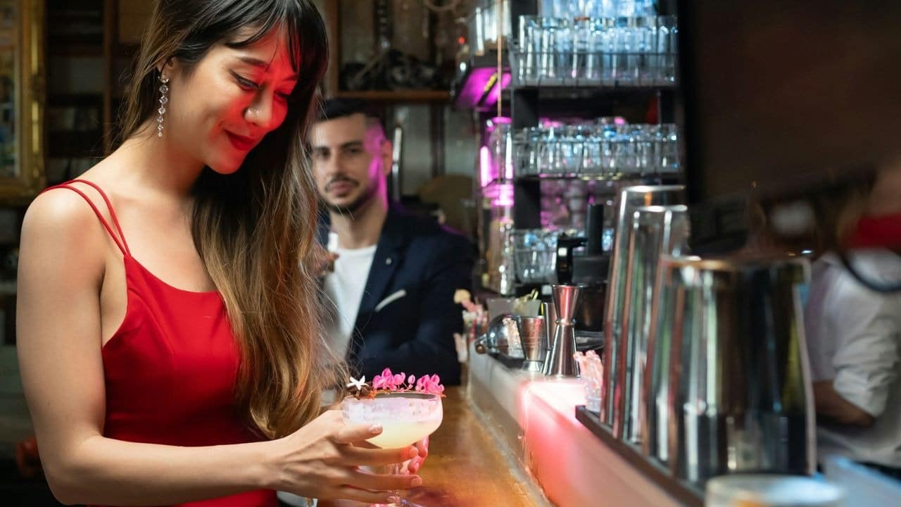 A woman in a red dress holds a cocktail at a dimly lit bar counter.
