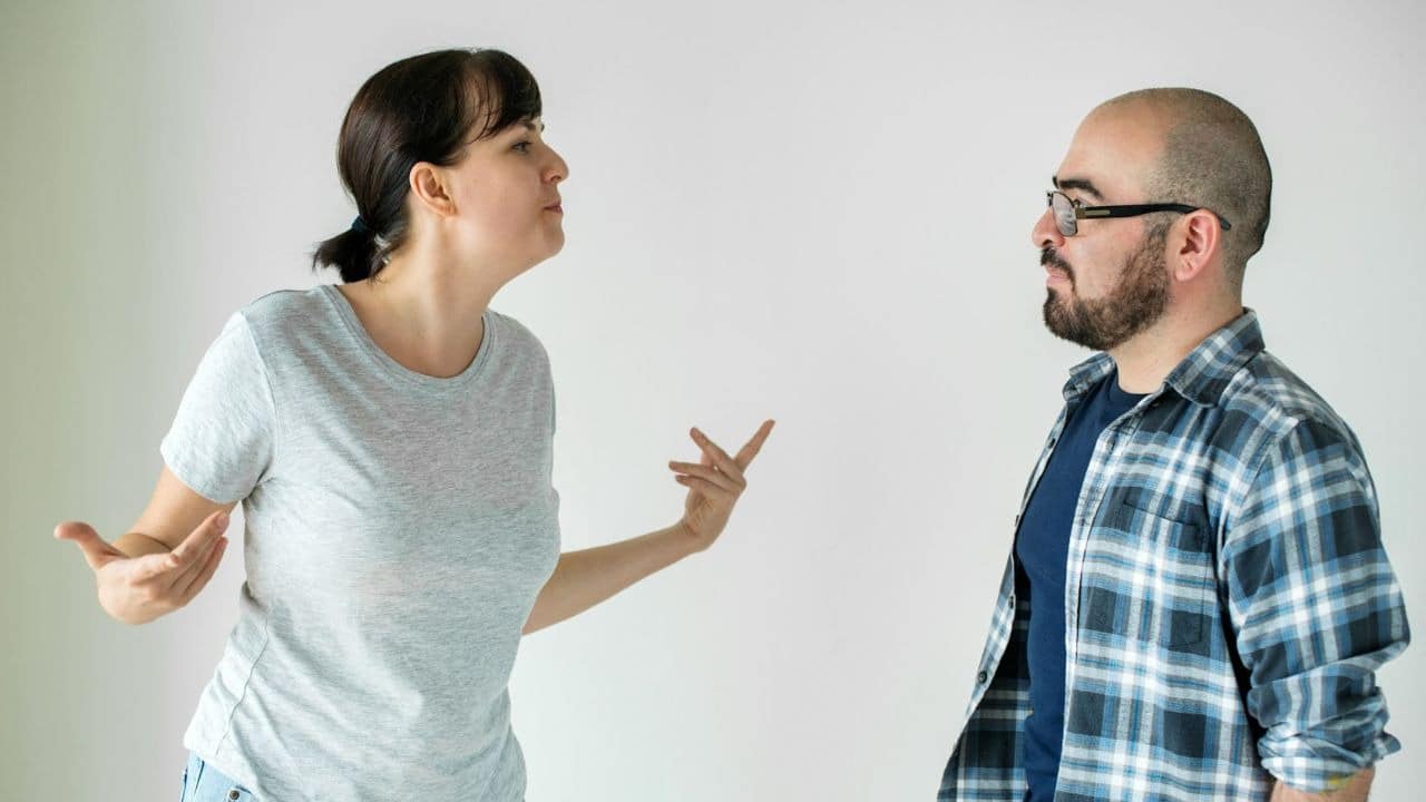 A woman in a grey shirt gestures broadly while facing a man in a plaid shirt.