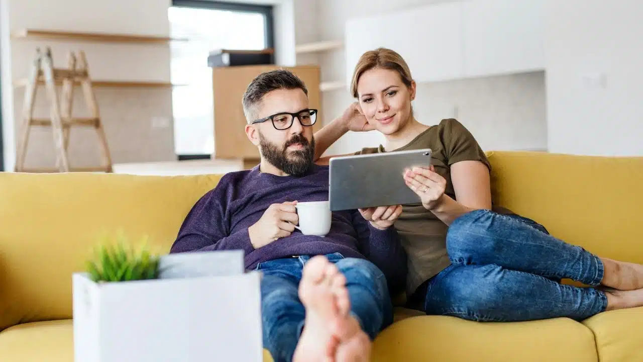 A man and woman sit on a yellow sofa looking at a silver tablet.