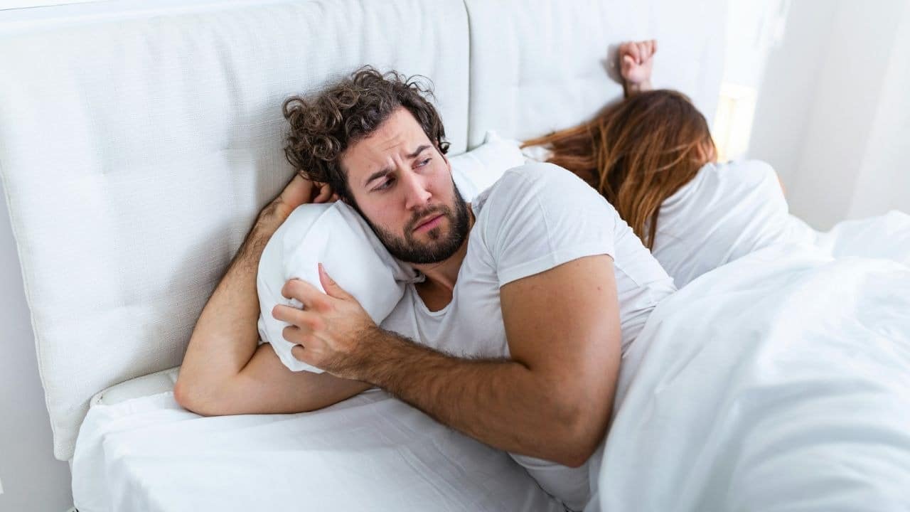 A man with curly hair lies in bed looking concerned while a woman sleeps.