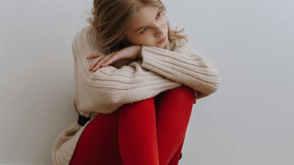 A woman in a beige sweater and red tights sits curled up, looking thoughtfully to the side.