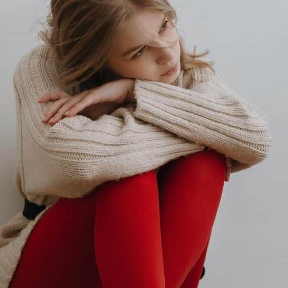 A woman in a beige sweater and red tights sits curled up, looking thoughtfully to the side.