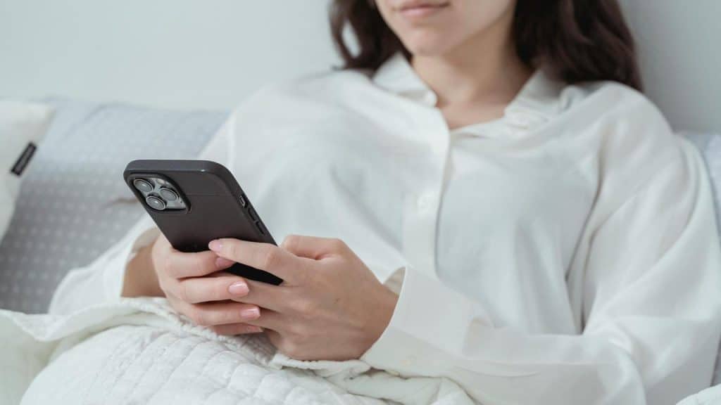 A woman in a white shirt lies in bed while using a smartphone.