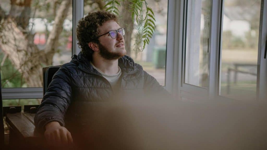 A man wearing glasses sits by a window, looking up thoughtfully.