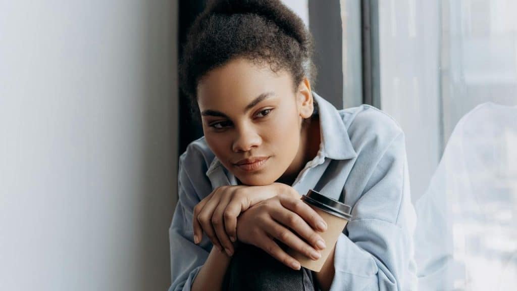 A woman sits by a window holding a coffee cup and looking thoughtful.
