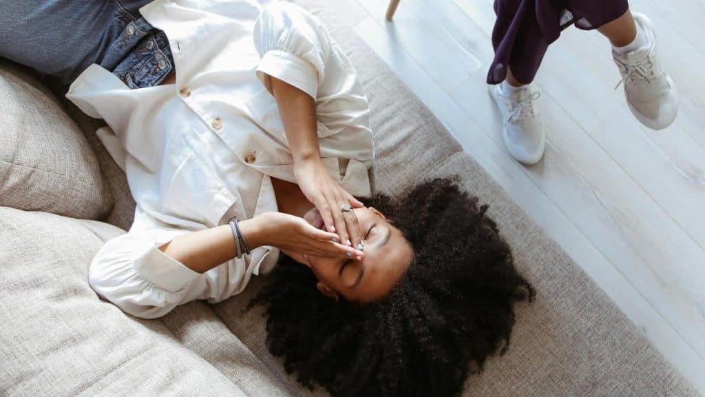 A woman lies on a couch covering her face while another person stands nearby.