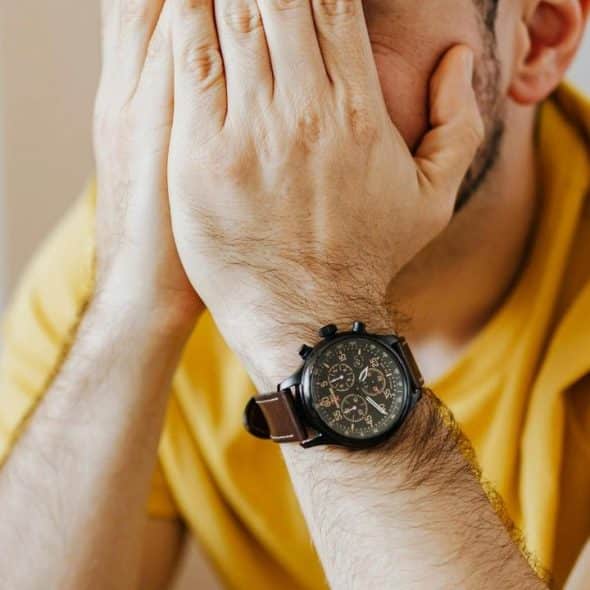 A person in a yellow shirt covering their face with their hands while wearing a wristwatch.
