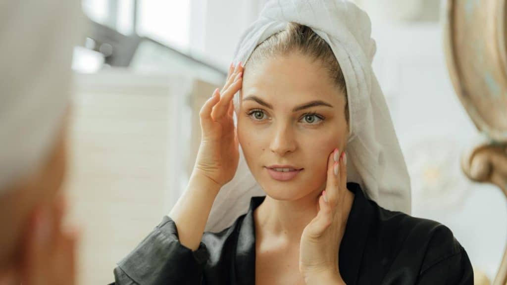 A woman with a towel on her head touching her face while looking in a mirror.
