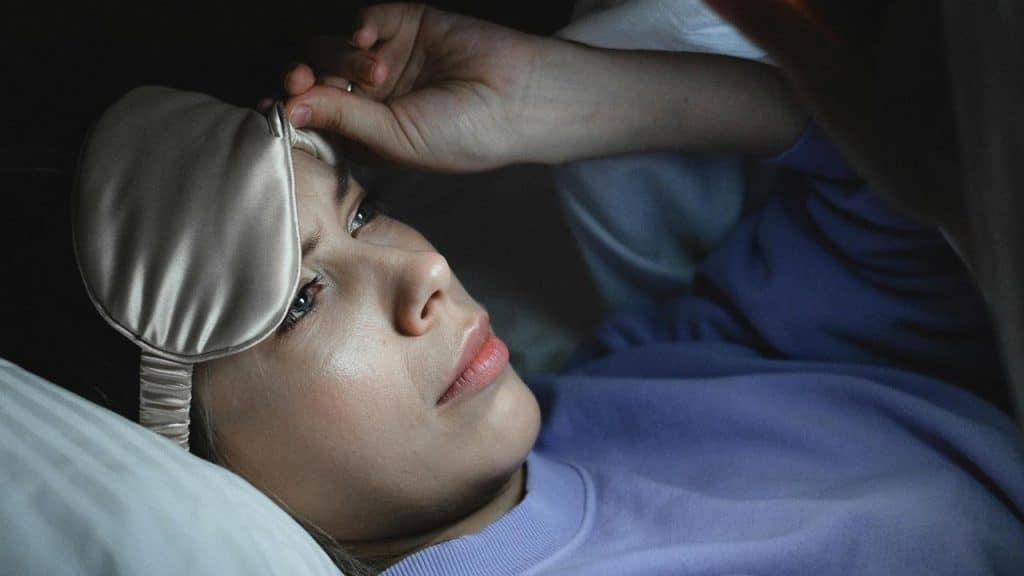 A woman lying awake in bed wearing a sleep mask pushed up on her forehead.
