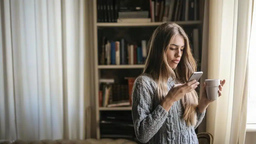 A woman holding a mug and looking at her phone indoors.