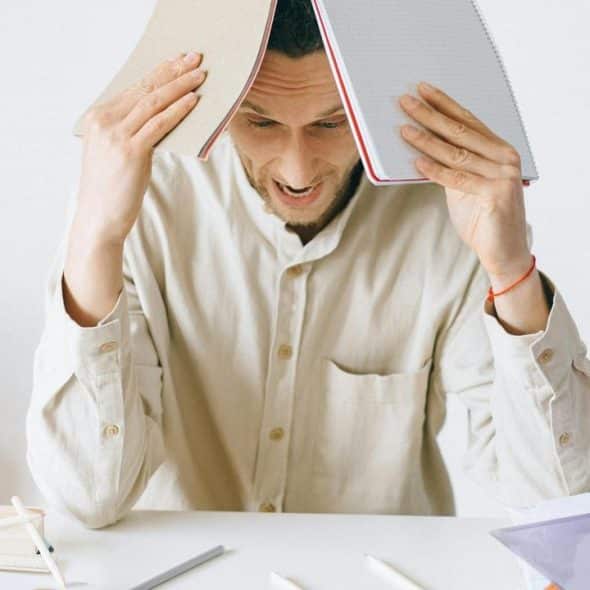 A stressed man holding two notebooks over his head at a desk.