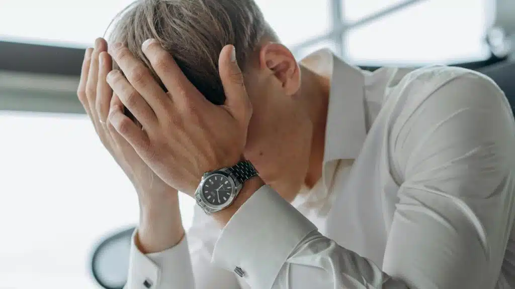 A stressed man in a white shirt covers his face with his hands.