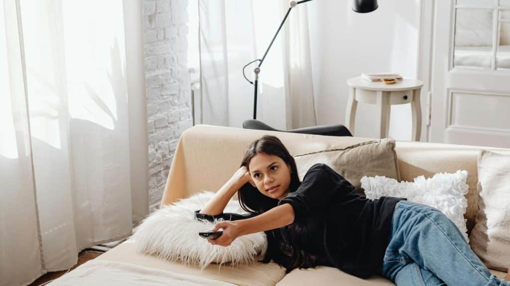 A woman lounges on a couch holding a TV remote in a bright living room.
