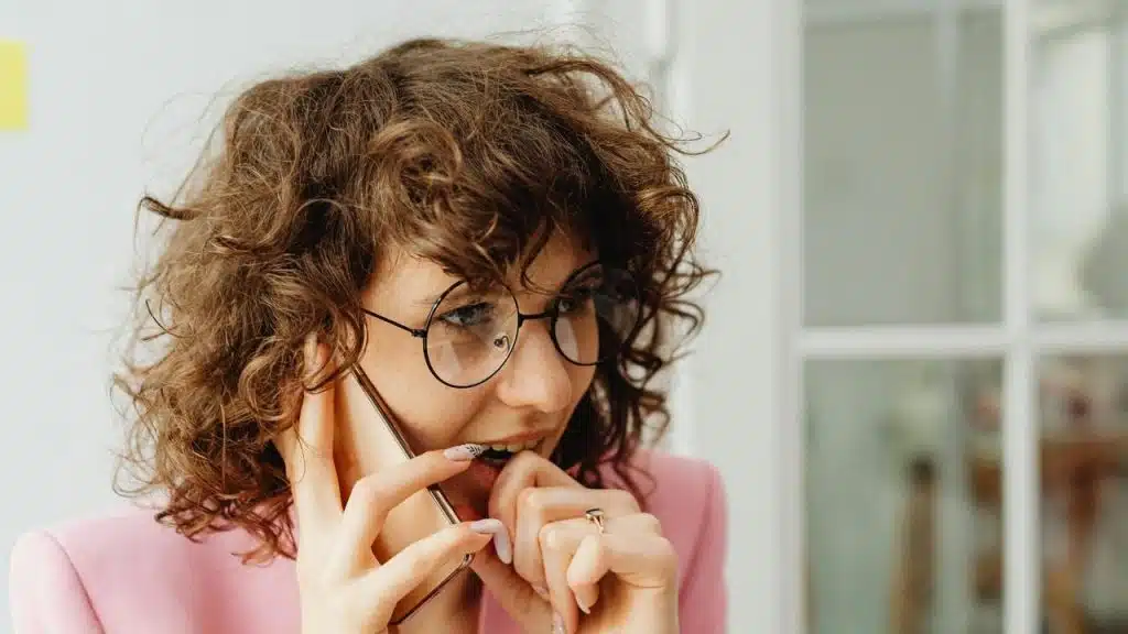 A worried woman with curly hair talks on the phone while biting her nails.