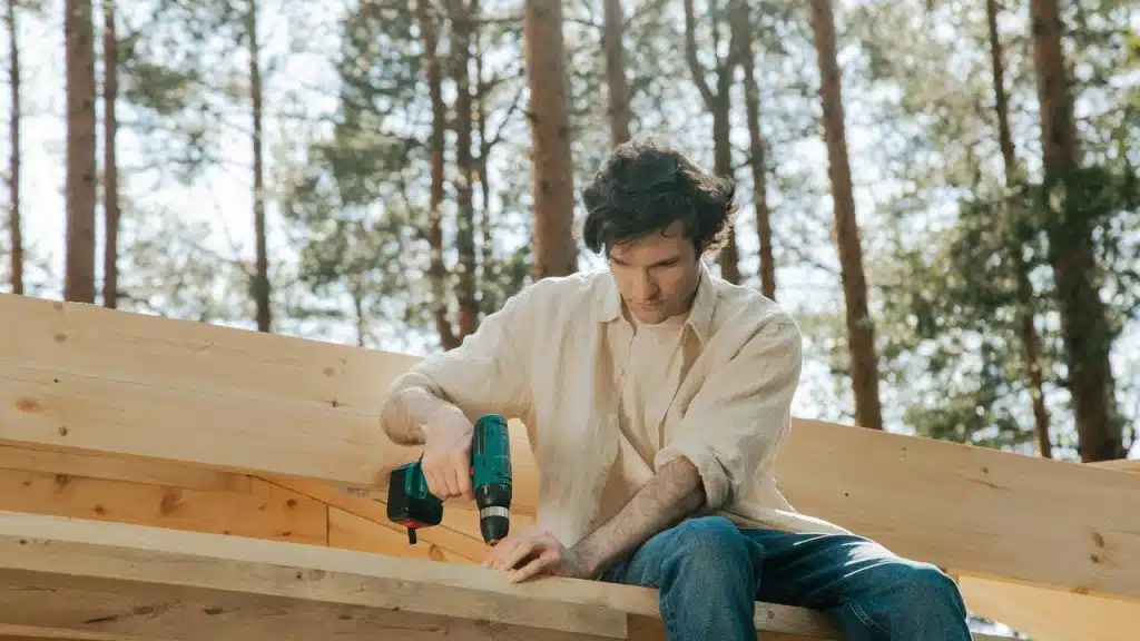 A man using a power drill while building a wooden structure outdoors.