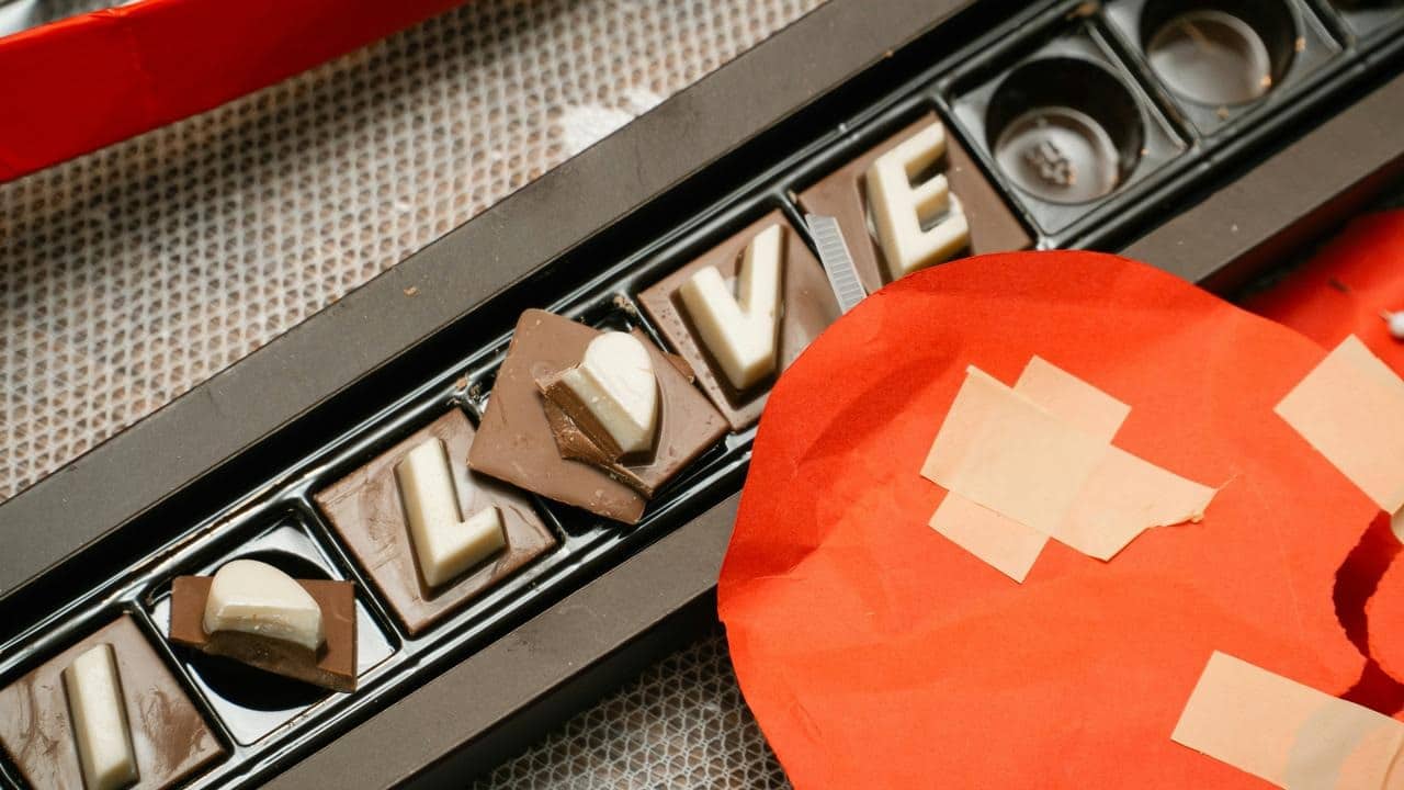 A box of chocolates spelling love beside a red heart decoration.