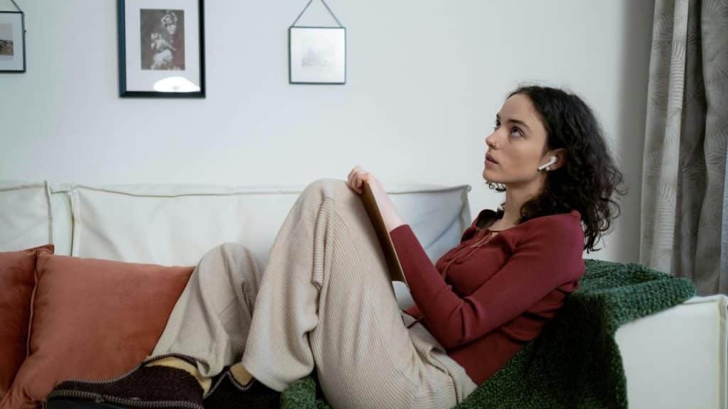 A woman sitting on a couch wearing earbuds and holding a notebook.