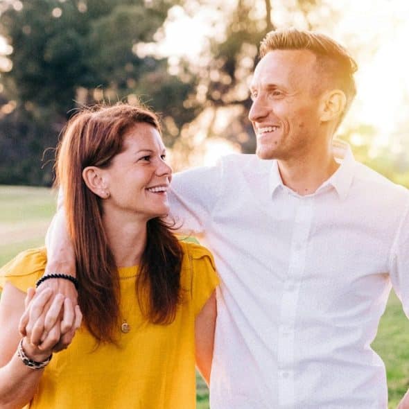A smiling couple with their arms around each other as they walk through a park.