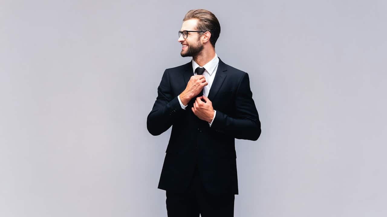 A bearded man wearing glasses and a suit smiles as he adjusts his tie.