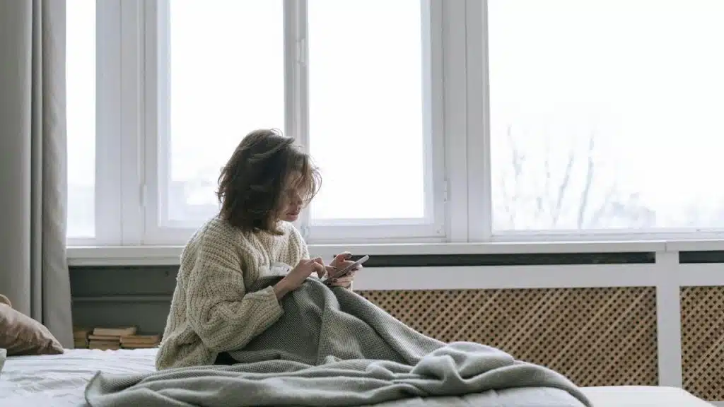 A woman sitting on a bed by a large window, wrapped in a blanket while using her phone.